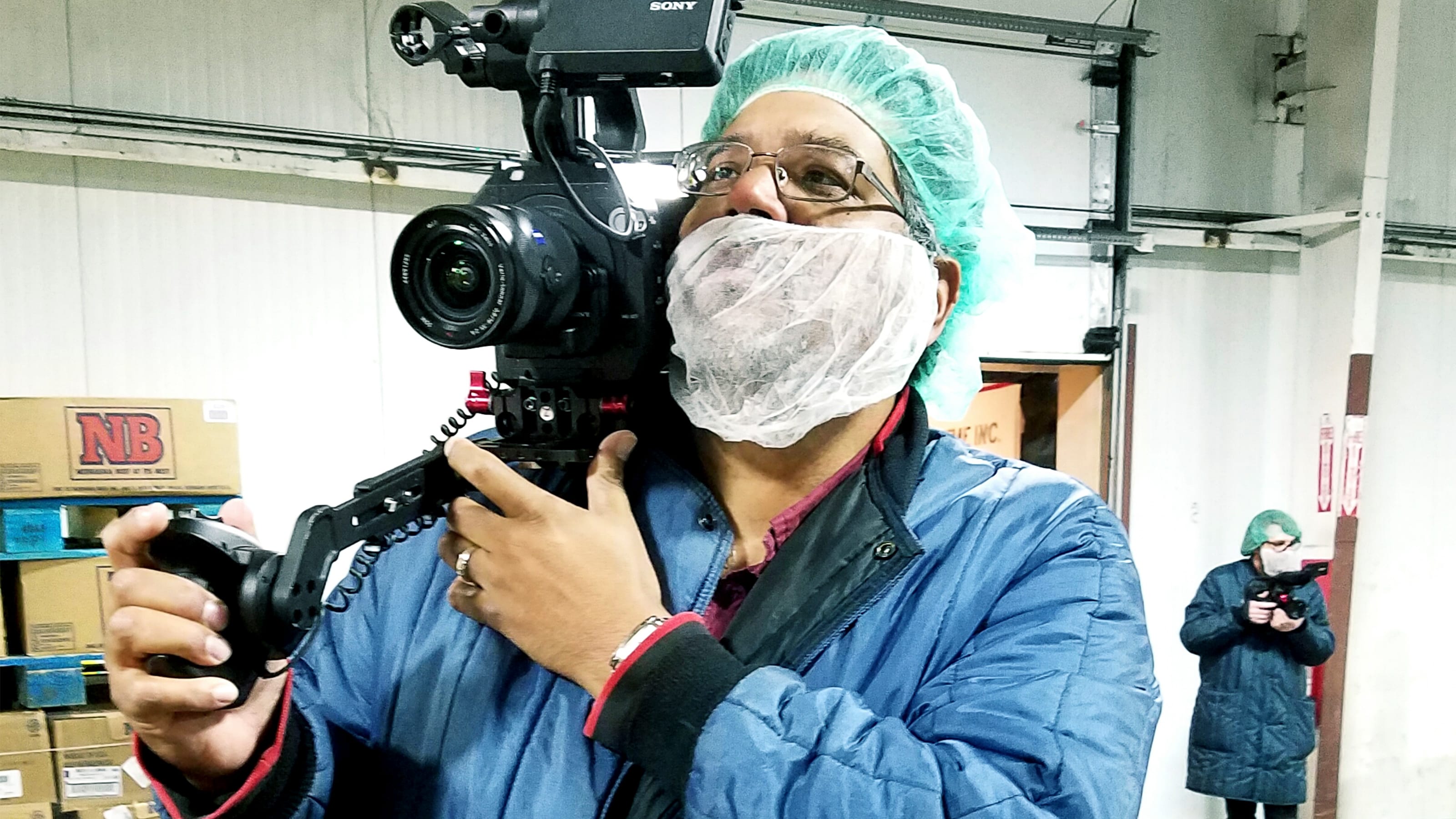 Man wearing a hairnet and beardnet operating a camera in a warehouse.