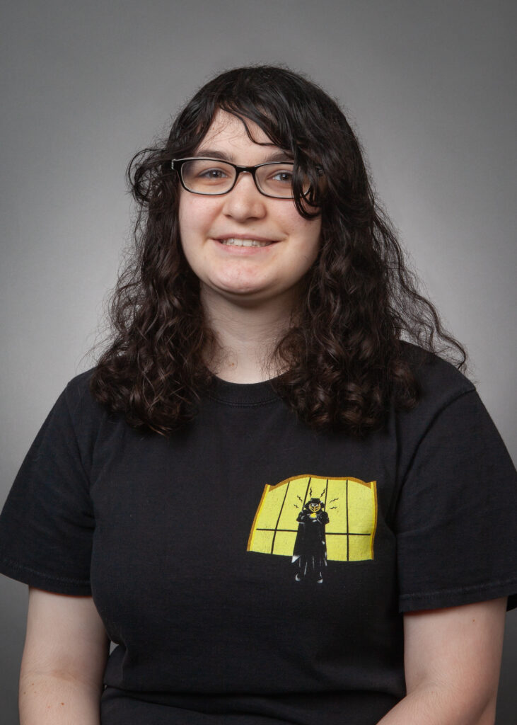 Headshot for Megan Carberry. Portrait of a young woman with long curly dark hair and glasses, wearing a black T-shirt with a yellow graphic design, set against a neutral gray background.